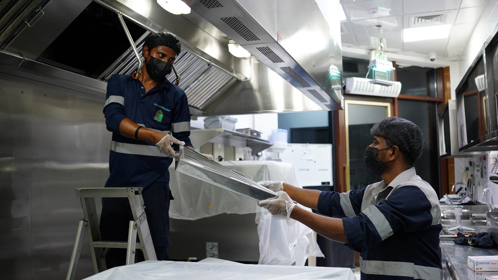 Two technicians in safety gear removing a stainless steel baffle filter from a commercial kitchen exhaust hood for deep cleaning